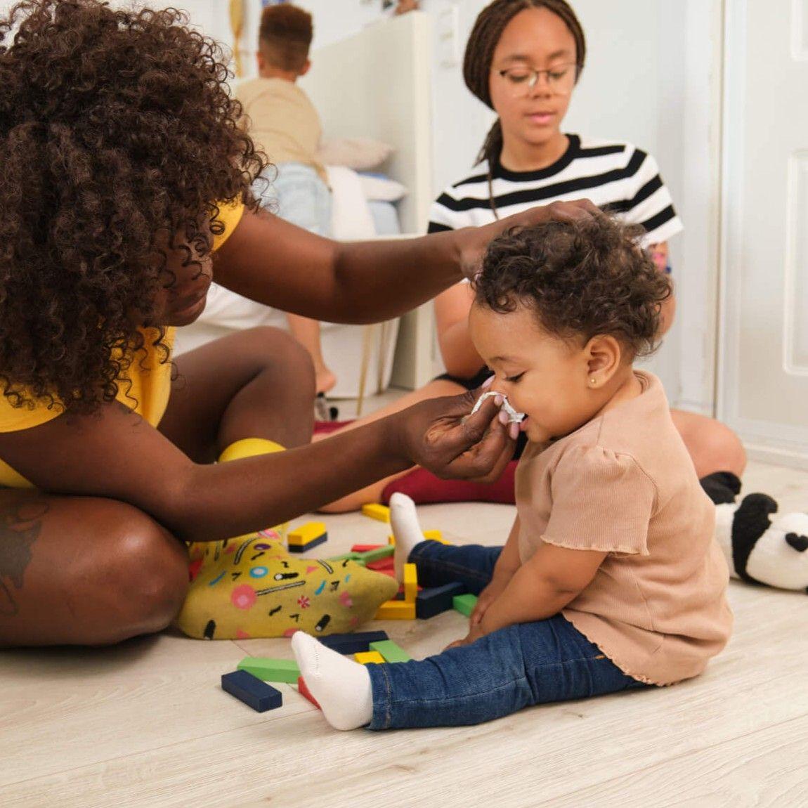 Caring mother wiping baby's nose in children's room with toys scattered around
