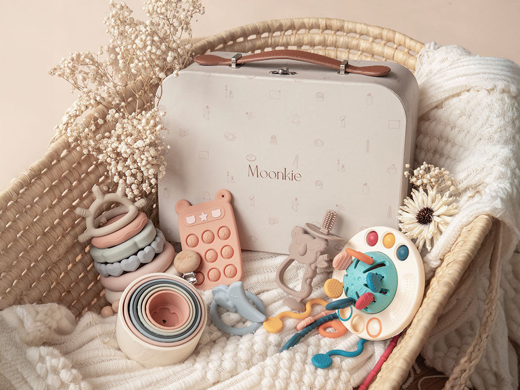 Collection of baby toys and a suitcase with 'Moonkie' branding on a soft fabric background.