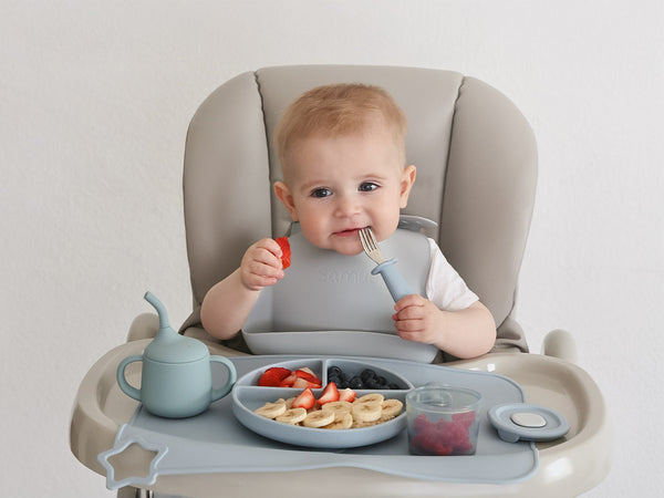 Baby in a high chair with a tray of food and utensils, eating fruits and vegetables.