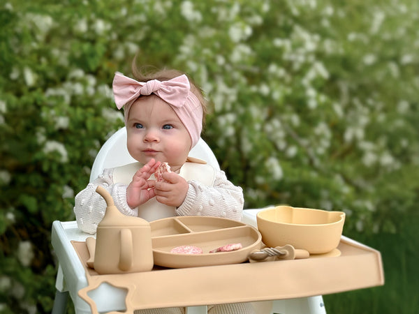 Baby sitting at a high chair with silicone  cutlery and a pink headband outdoors.