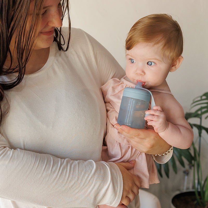 Woman holding a baby who is drinking from a sippy cup indoors.